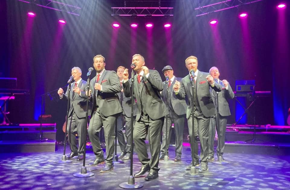 A male vocal ensemble performing on stage with microphones under pink and blue stage lighting.