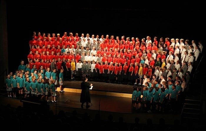 A diverse choir, organized by shirt color into sections, performs on a dark stage led by a conductor in black.